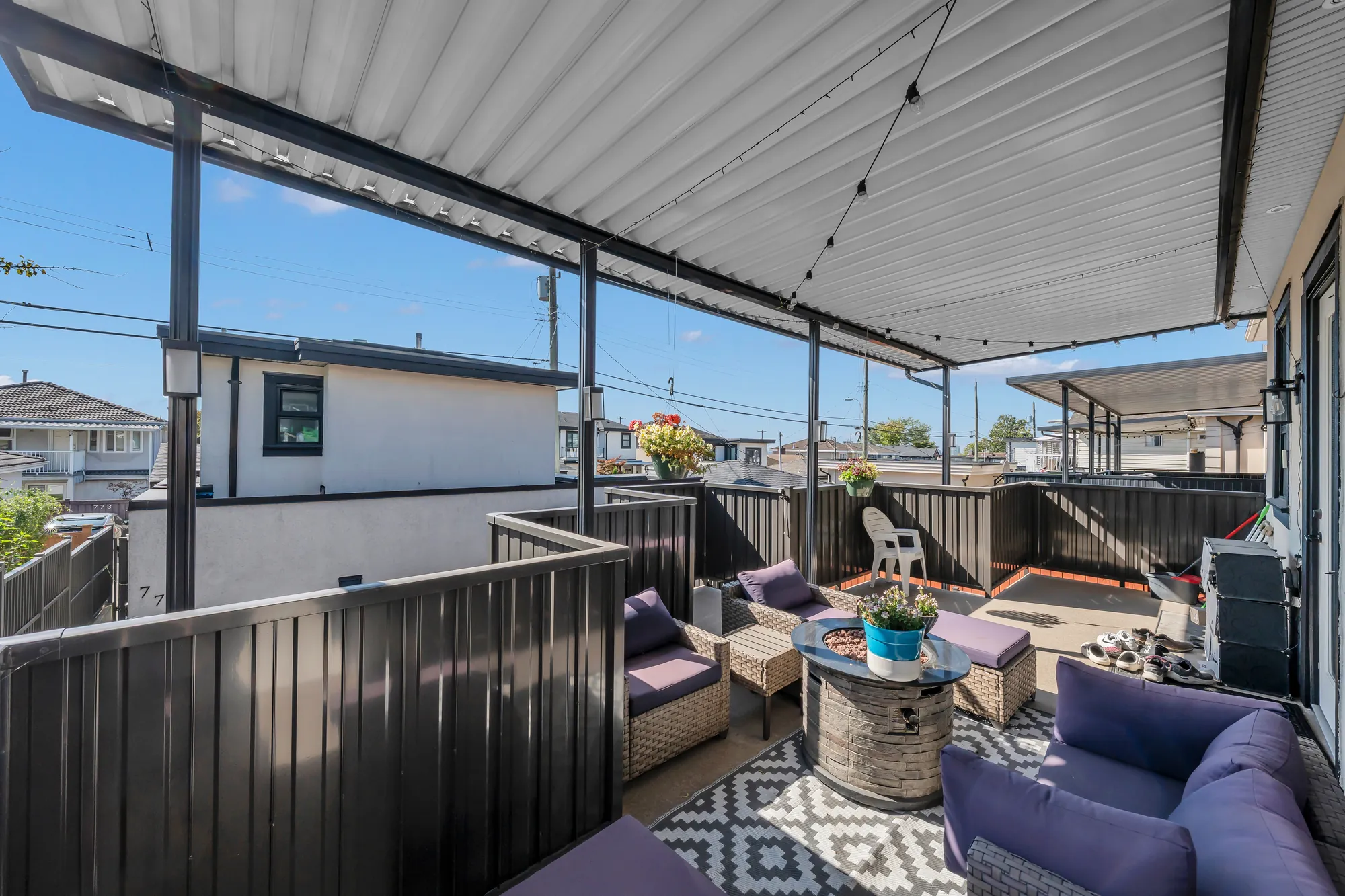 The covered balcony over the carport: a wicker sofa set with purple cushions around a fire table on a patterned rug, a metal roof on posts, the neighbours' roofs beyond the railing