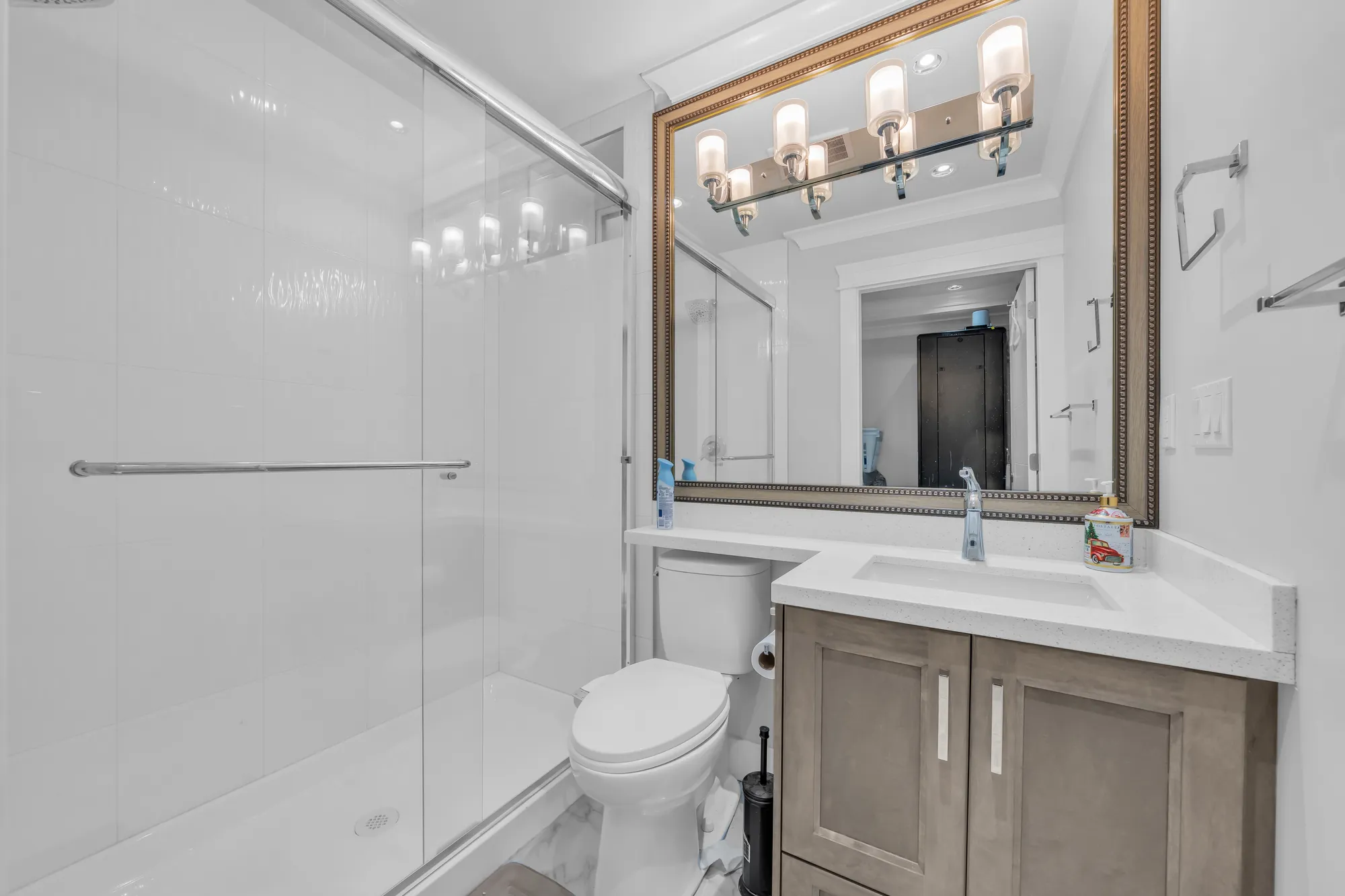 A bathroom with a glass shower, a wood vanity with a white counter, a gold-framed mirror under a light bar