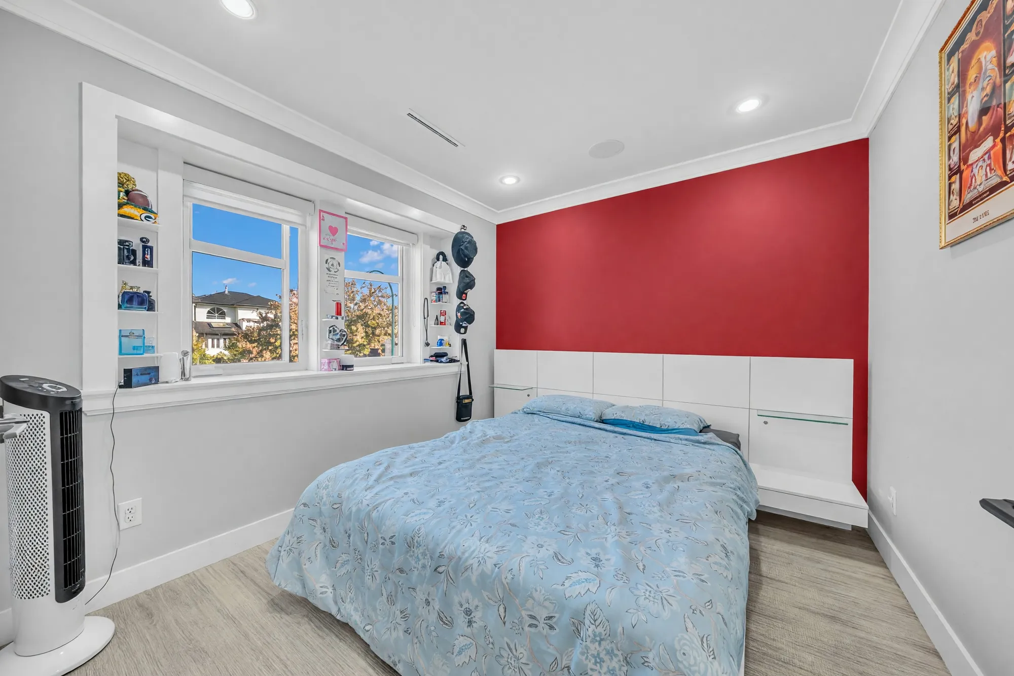 A bedroom with a red feature wall, a bed under a white headboard shelf, two windows, a white fan