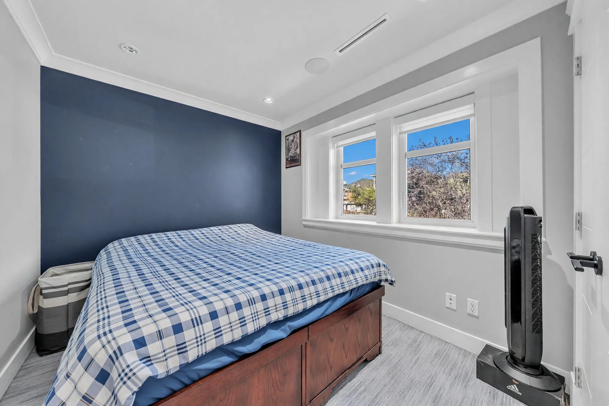 A bedroom with a navy feature wall, a wooden bed with a plaid cover, a window, a tower fan