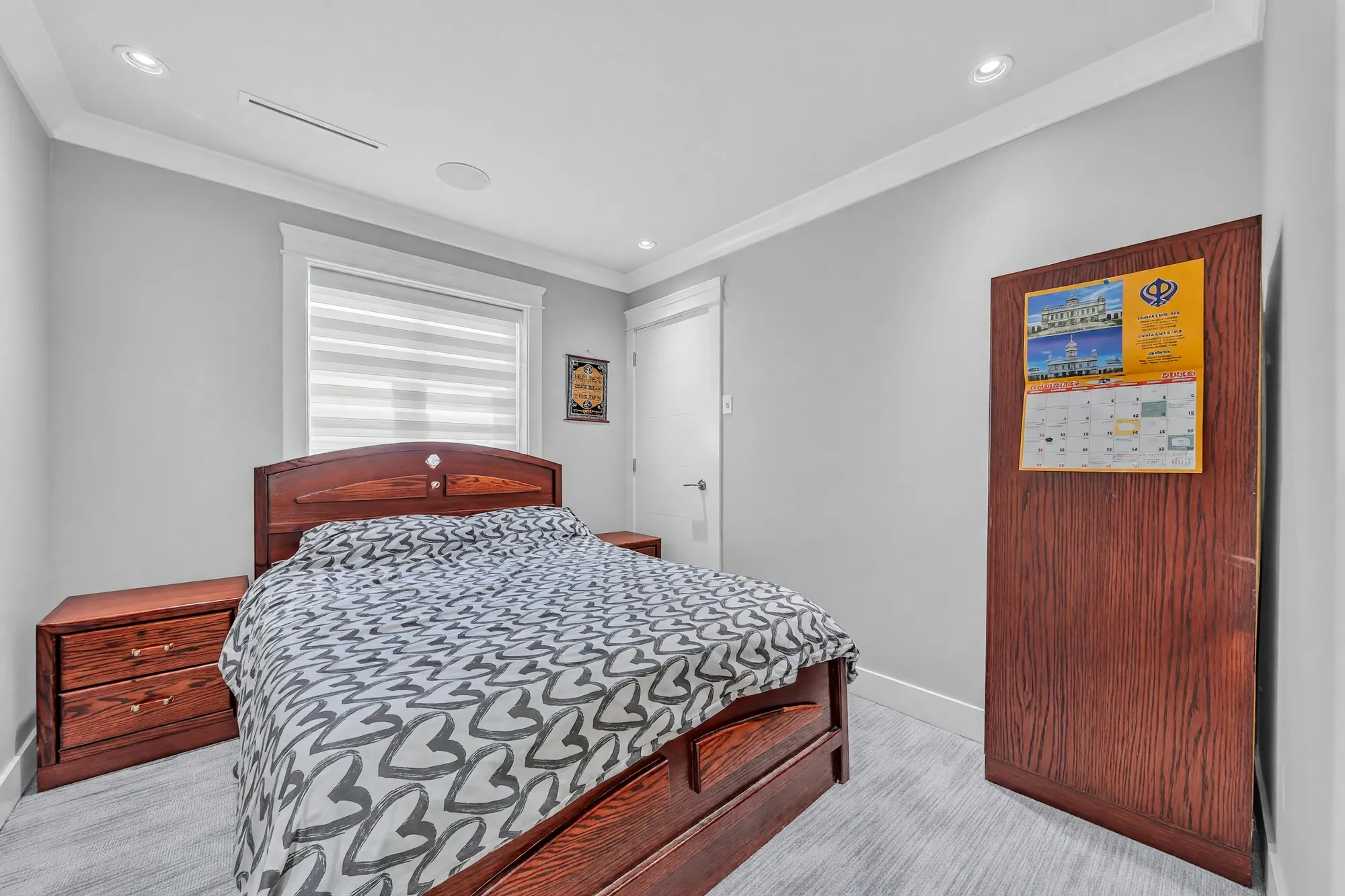 A bedroom with a wooden bed, a dresser, a window with a blind, a wardrobe with a calendar