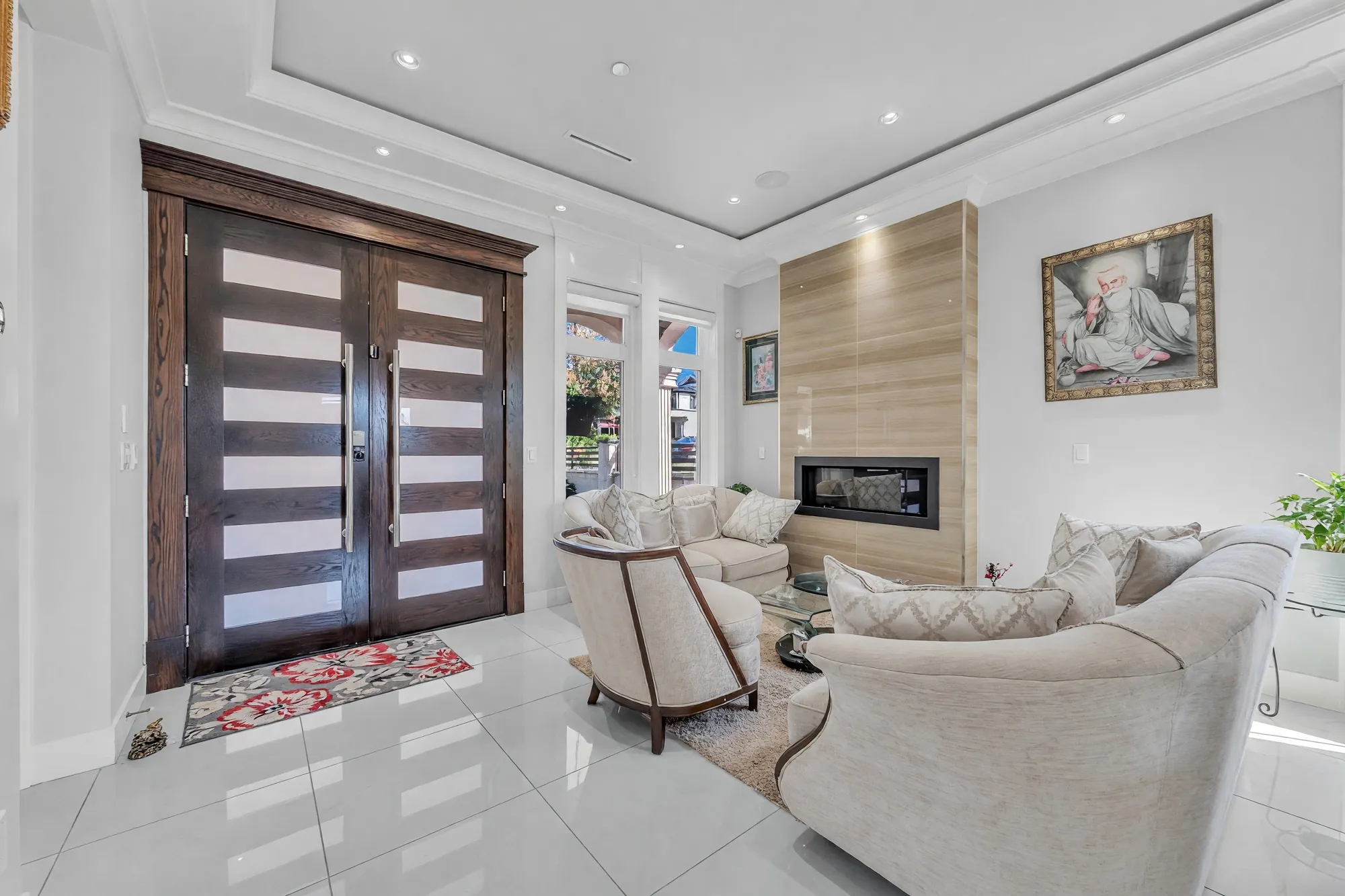 The foyer and living room from the front door: double doors in dark wood with glass slats, cream sofas with wood trim, the fireplace in its wall of pale wood, a painting