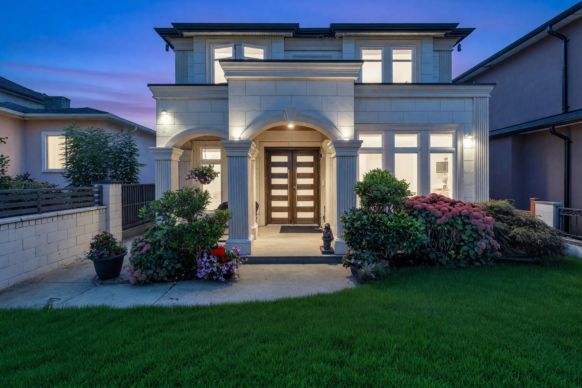 768 East 58th Avenue at dusk: a two-storey house in cream limestone with an arched portico on square columns, the windows lit, the lawn and the shrubs in front