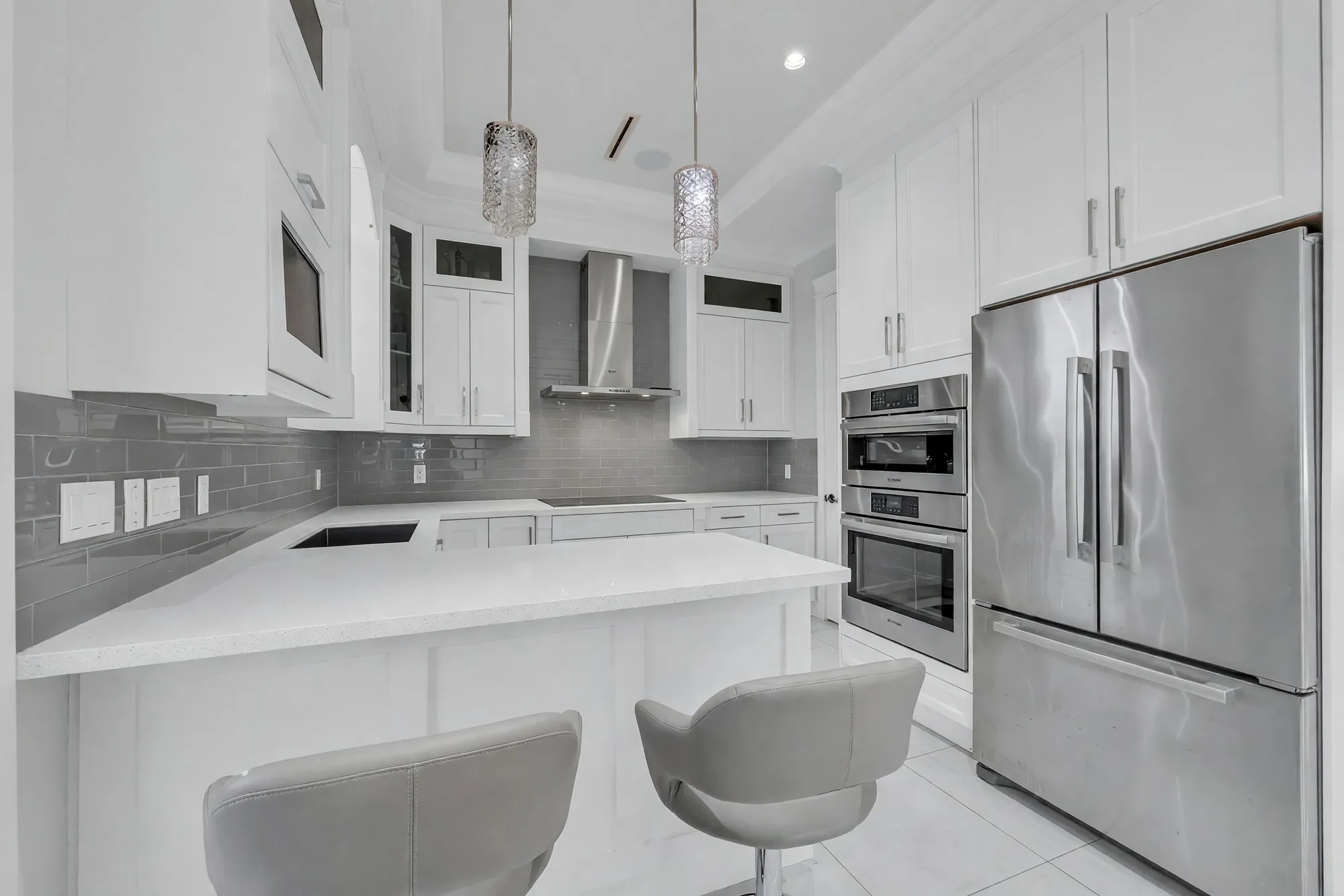The kitchen: white shaker cabinets, a quartz island with grey stools, a stainless refrigerator and wall ovens, a grey tile backsplash, crystal pendants
