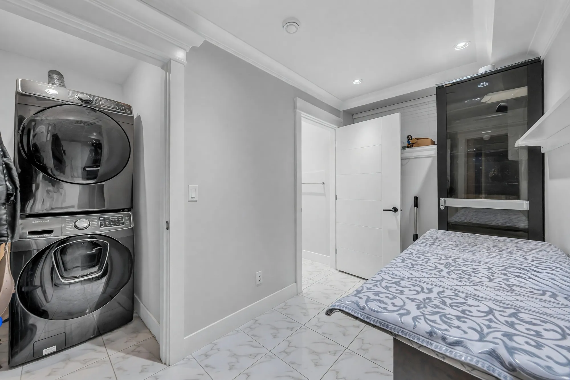 A stacked washer and dryer beside a hall with a bed and a red cabinet, marble-look tile