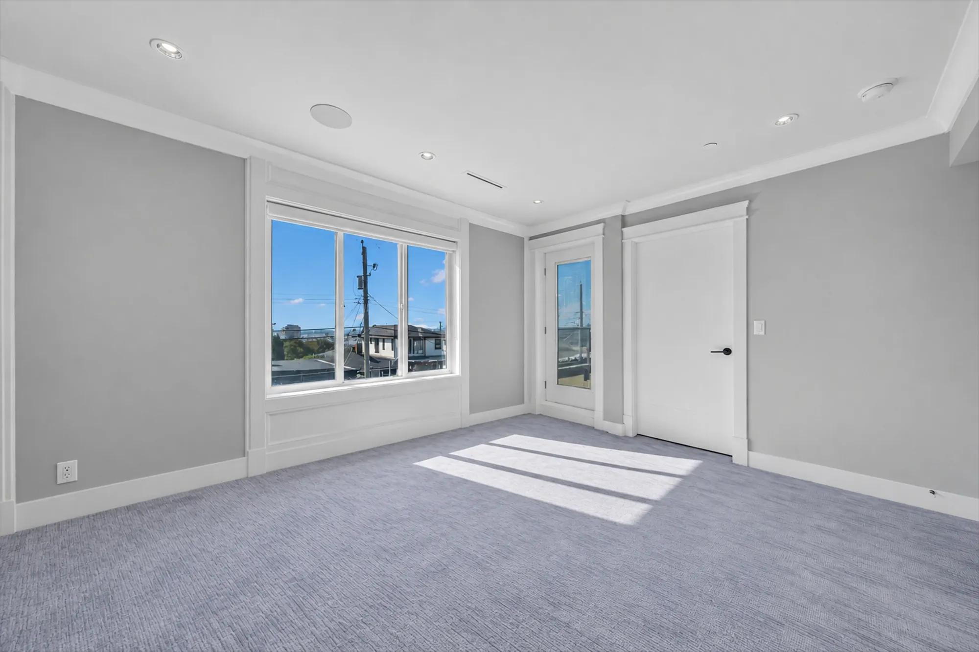 The primary bedroom shown empty: grey carpet, a window to the street, the door to the balcony and the door to the bathroom