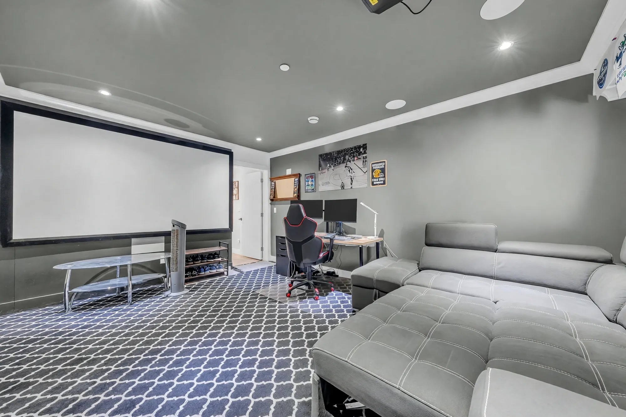 The sitting area from the other end: a grey leather sectional facing a projector screen, the desk beyond